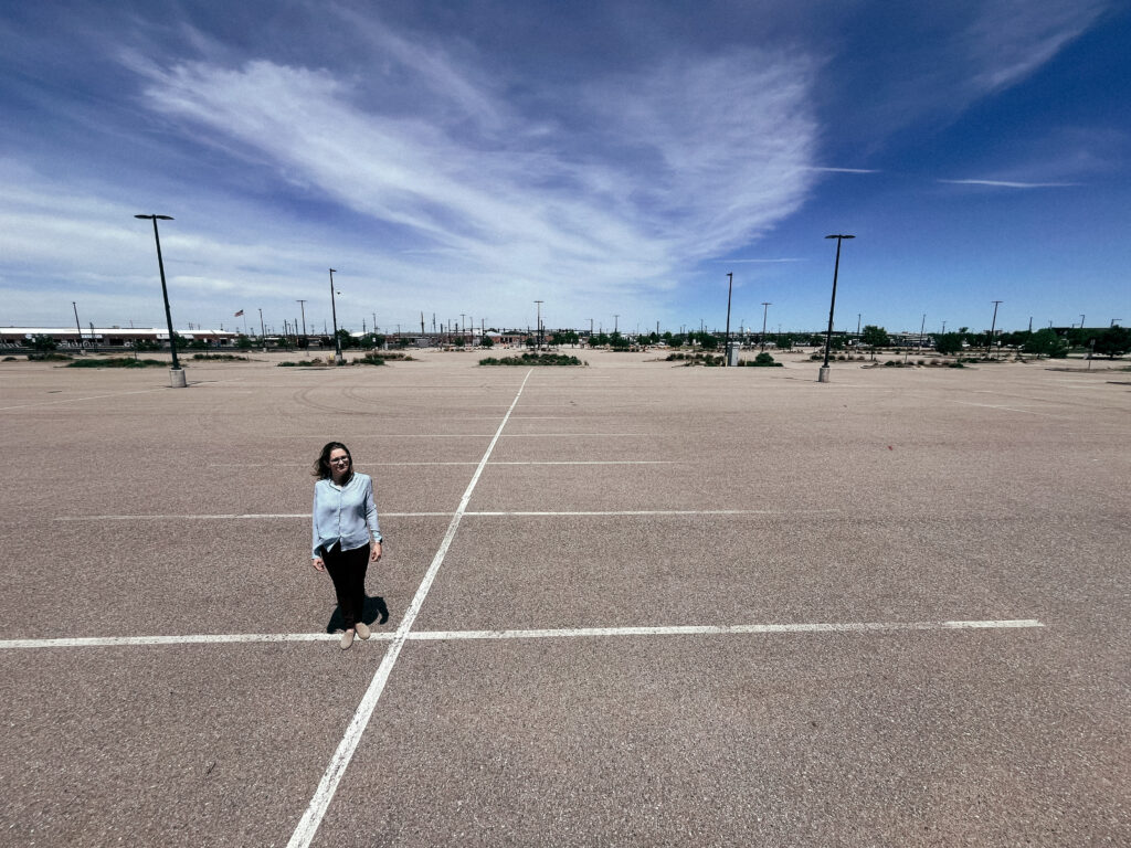A woman wearing a light colored long sleeve shirt and black pants stands in a parking lot.