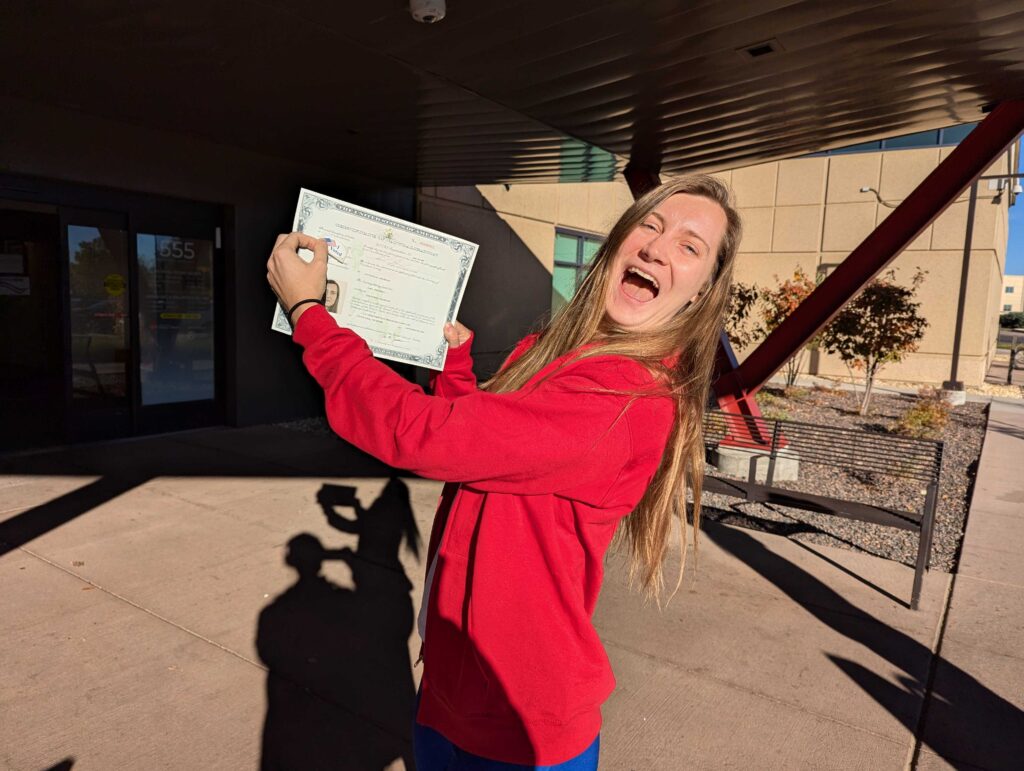 Woman in red jacket voting for the first time in her life, proudly displays her naturalization certificate and smiles broadly while voting in Lakewood, Colorado.