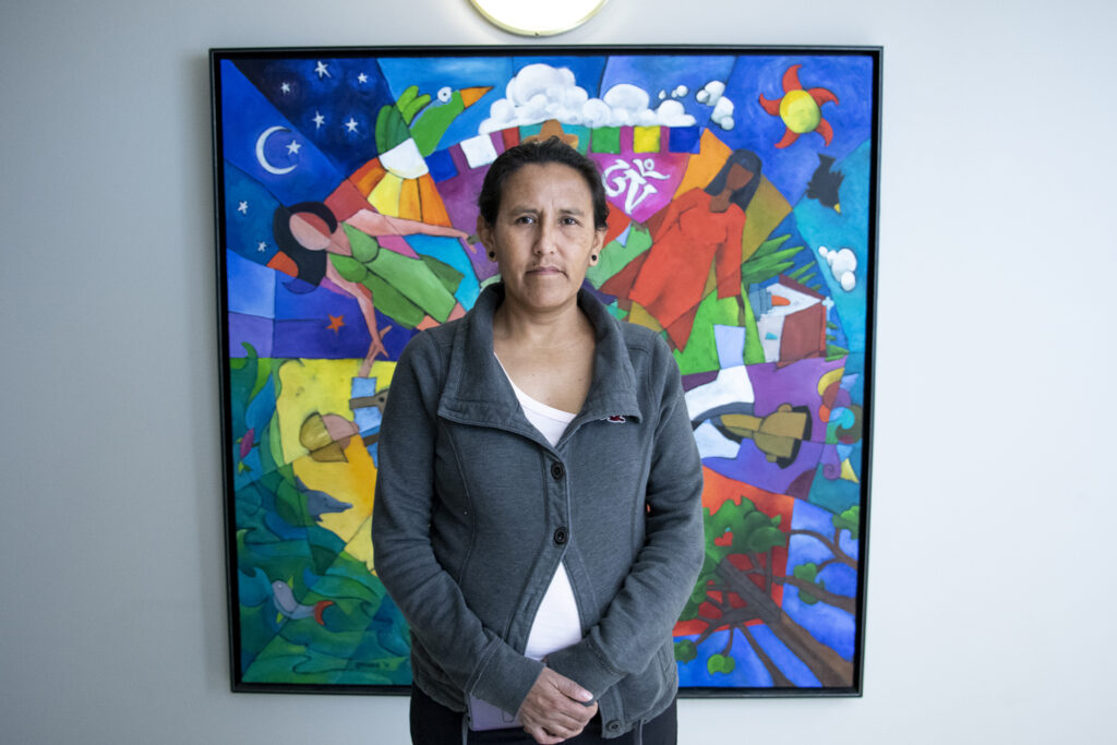 A woman in a gray sweater stands in front of a colorful painting with her hands folded.