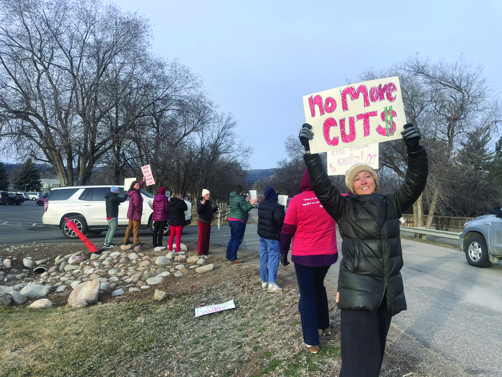 A woman in a black puffy jacket holds up a sigh that says "no more cuts" as about half a dozen other also hold signs as car drive past them.