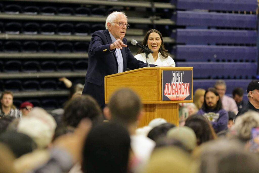 Bernie Sanders and Alexandria Ocasio-Cortez stand together at a podium reading "Fight Oligarchy" from a podium that reads "Fight Oligarchy," surrounded by a large crowd.
