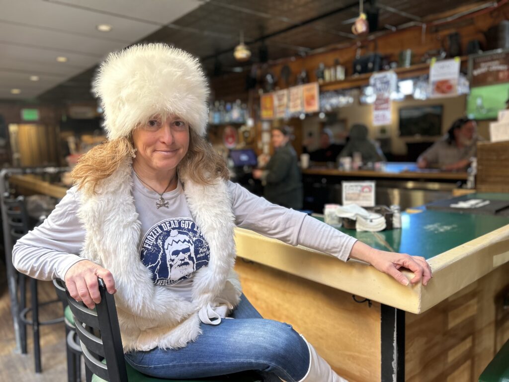 Stephanie Andelman sits at a bar in Nederland, Colorado wearing a "Frozen Dead Guy Day" T-shirt and a white faux fur hat and vest.