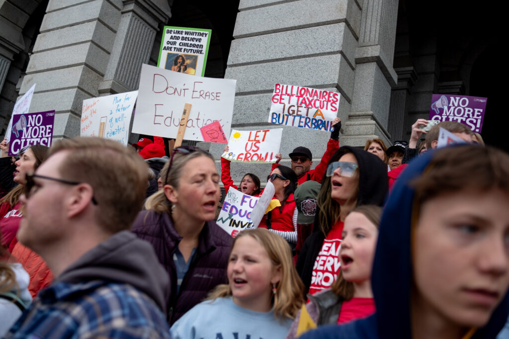 The Colorado state Capitol building's west steps are seen crowded with people holding signs that say "No more cuts."