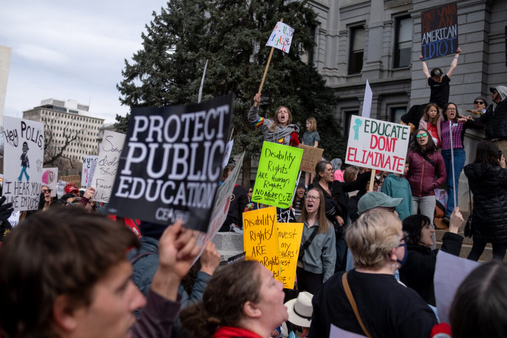 A crowd gathers on the steps of the Capitl with signs that say "Educate don't eliminate" and "protect public education."