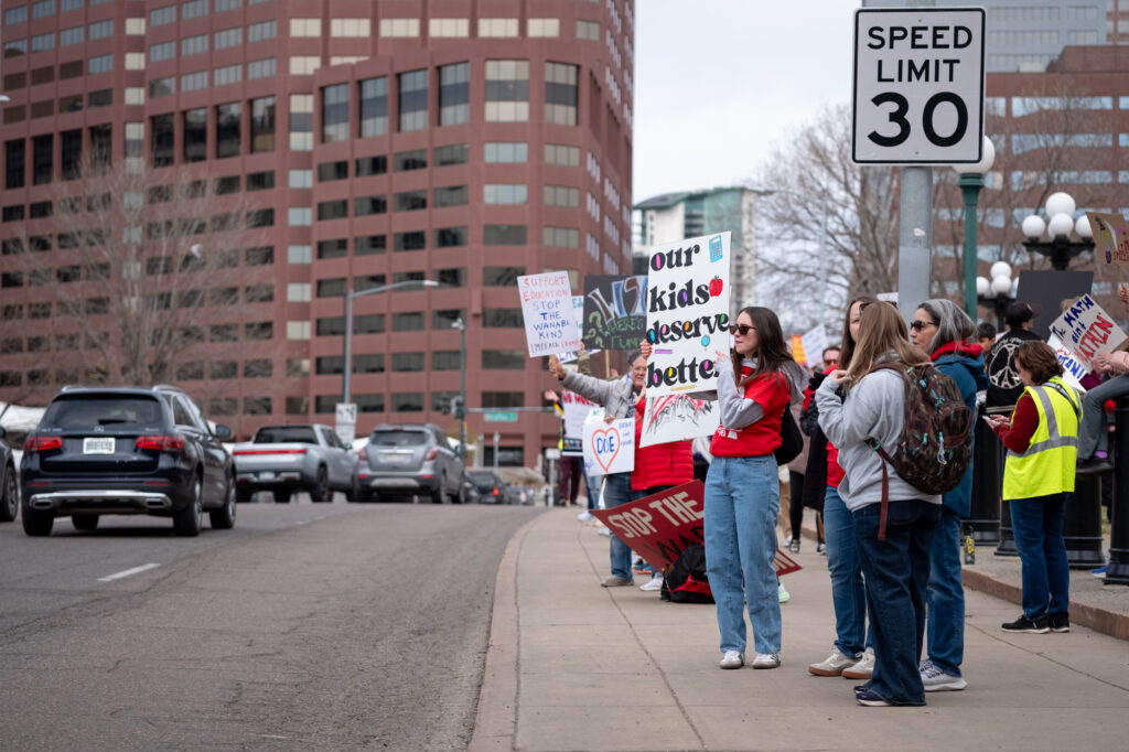 Protester line the sidewalks along Lincoln Street in front of the Capitol. Cars are driving by as people hold up signs, one that says "our kids deserve better."