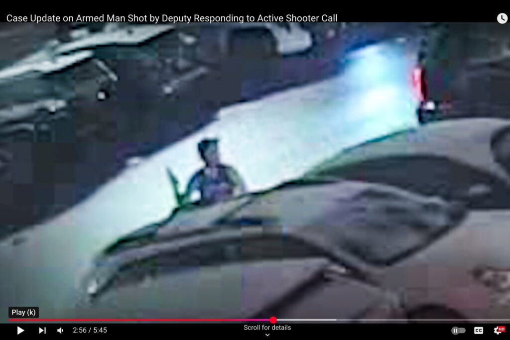 Man in tank top jersey holding a gun in his right hand and standing between two cars