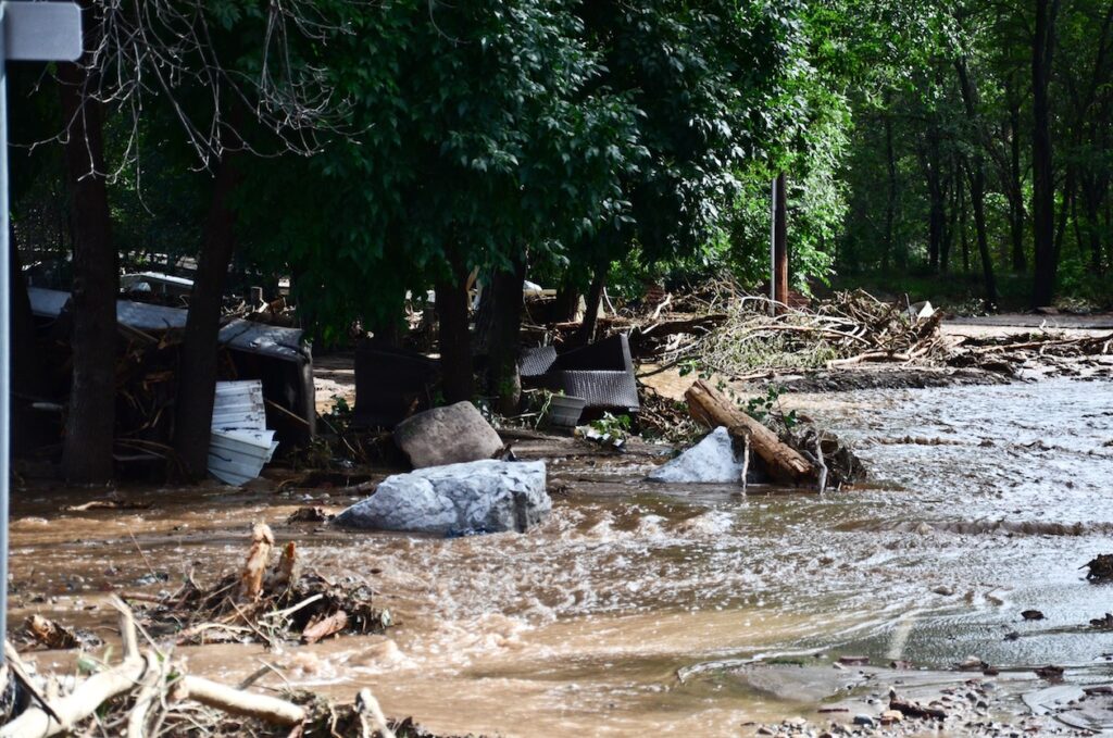 Flood water runs down a street with a lot of debris in it. There are trees on the left side and a lot of wood.