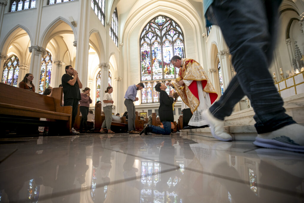 A priest in a robe administers the host to a kneeling person in a large church.