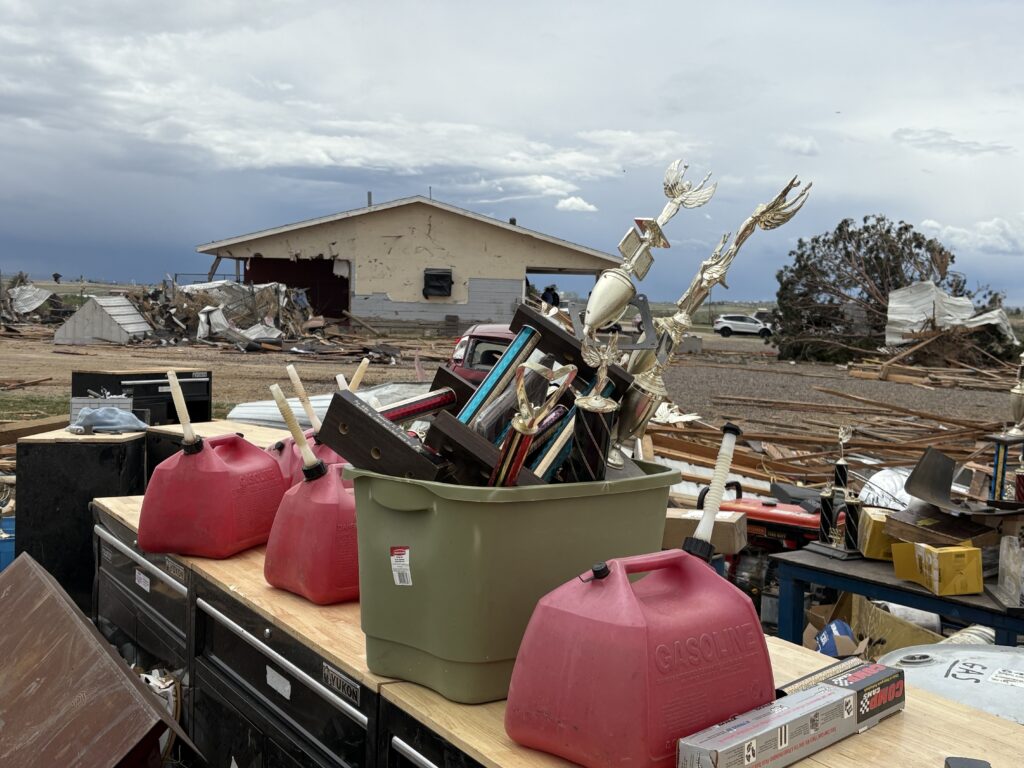 Golden colored trophies dumped in a bin on a table amongst debris. A wrecked house sits in the background.