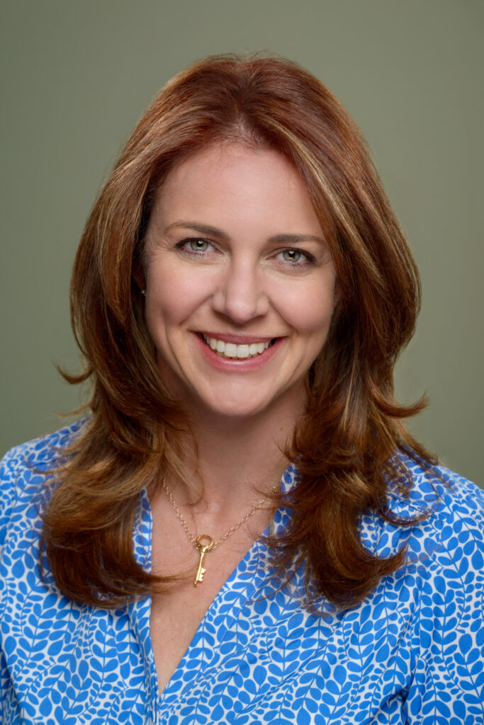 woman with long strawberry blonde hair smiling at camera wearing blue and white print shirt