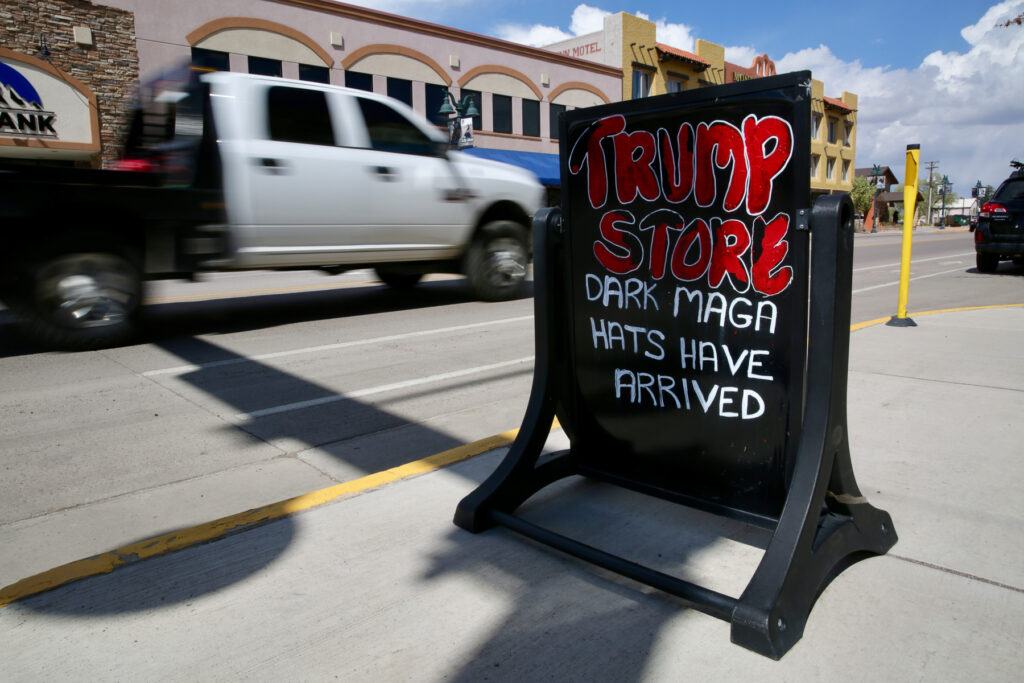 A sandwich board sign reading “Dark MAGA hats have arrived” is displayed on a sidewalk while a white pickup truck drives past.