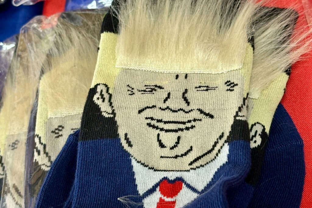 A pair of socks featuring the likeness of Donald Trump with fluffy blonde hair is shown in a close-up.