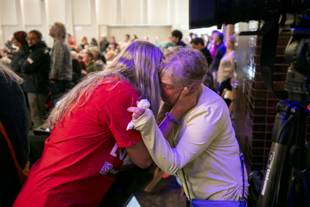 Two women are embracing each other in a crowded room, with one woman holding a tissue. They are surrounded by other people