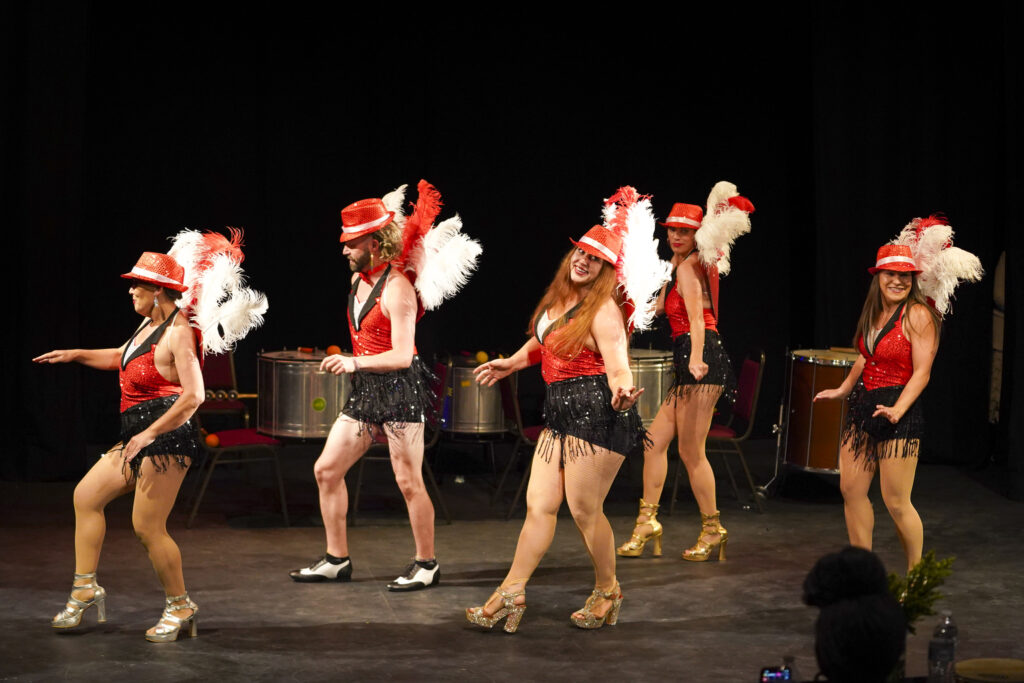 Five dancers in black shorts with fringe, red halter tops and red hats, plus large red and white feathers off their backs, perform for the competition’s judges.