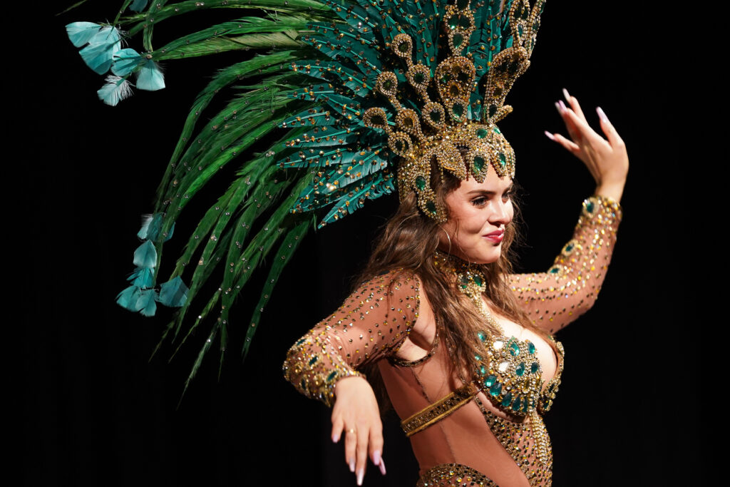 A dancer twists her body in a samba-style dance move. She wears a large headdress with peacock-like feathers and a nude-colored, long-sleeved leotard that is bedazzled with teal and gold gems.