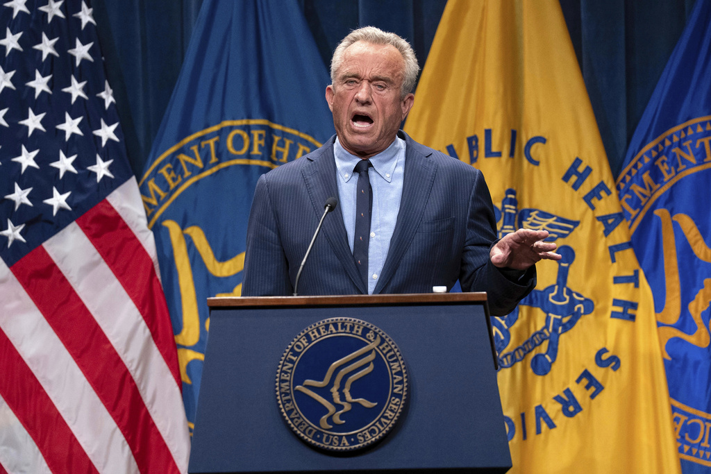 A man in a dark suit and tie stands behind a podium with a microphone, giving a speech. The podium is has multiple flags and the words "United States Army" are displayed.