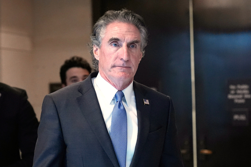 A man in a dark suit and blue tie is standing in a room with a black door. He has gray hair and is looking off to the side.