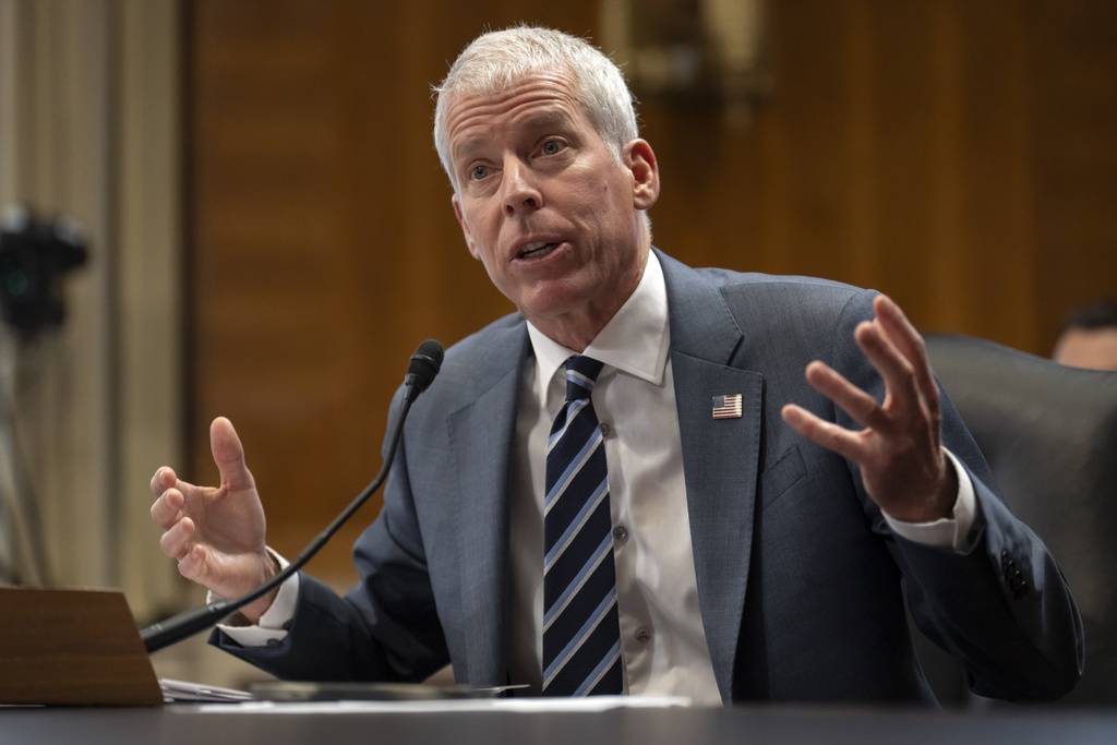 A man in a gray suit and blue tie is seated at a desk, gesturing with his hands. He is speaking into a microphone placed on the desk. The background features a wooden panel and a camera