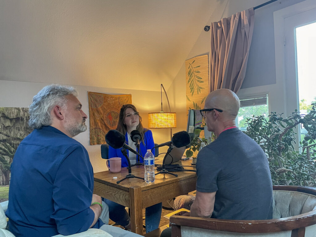 Woman in blue jacket interview two men at wooden table surrounded by plants and art.