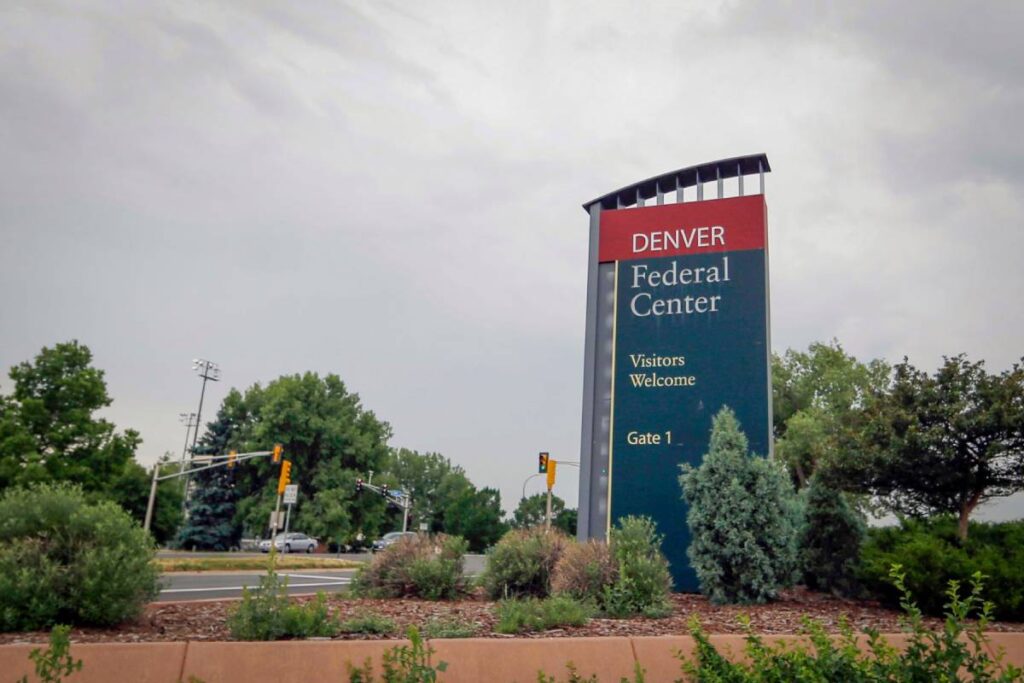 The image features a large sign with the words "Denver Federal Center" written on it. The sign is located in front of a building, and there are trees in the background. The scene is set on a street, and there are cars parked nearby.
