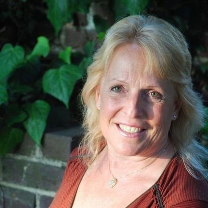 woman smiling with blonde hair and brown shirt in front of brick wall with plants