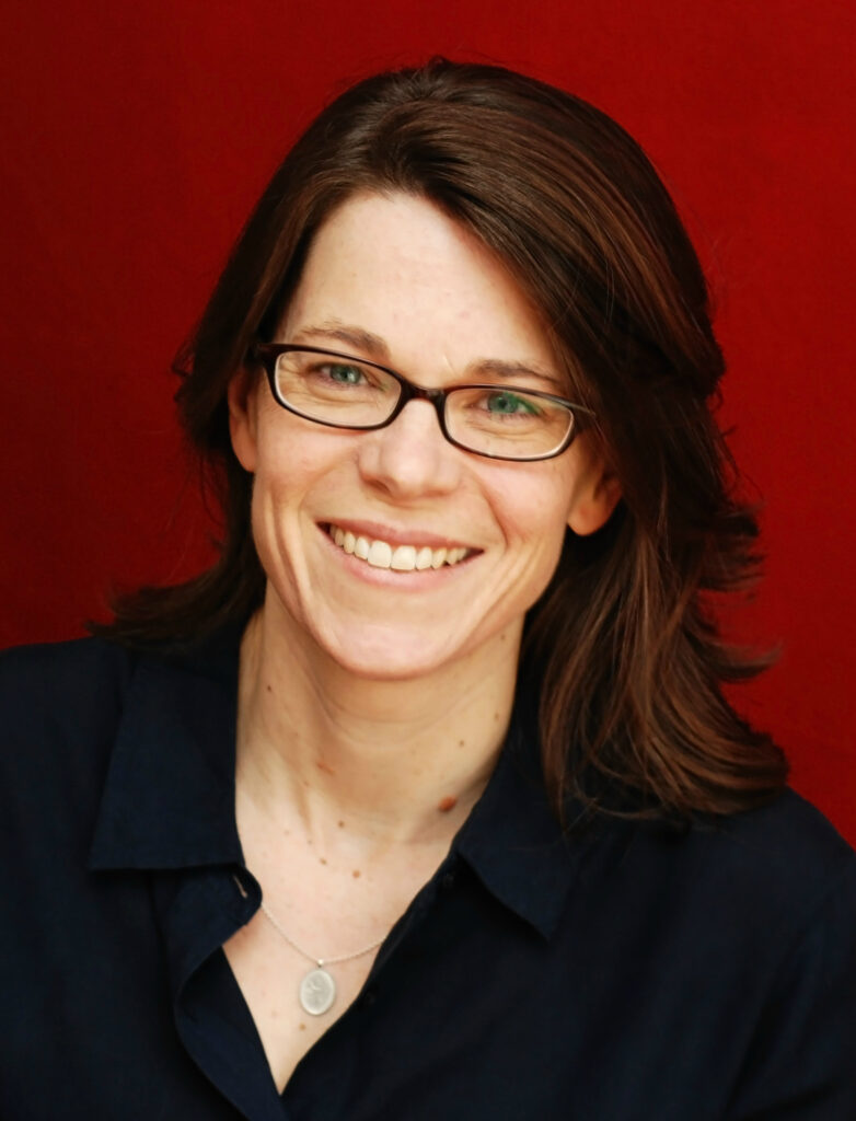 Susan Schulten in front of a red background, smiling and wearing glasses with a navy blue shirt.