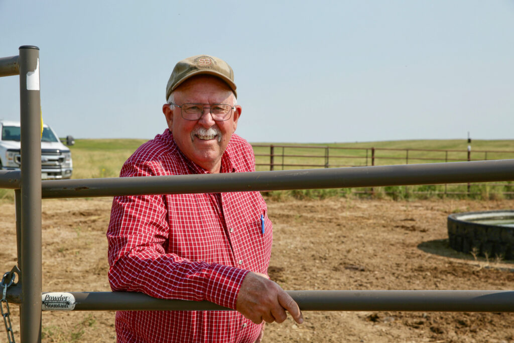 A man in a red plaid button-up shirt leans against a gate in the eastern Colorado plains.