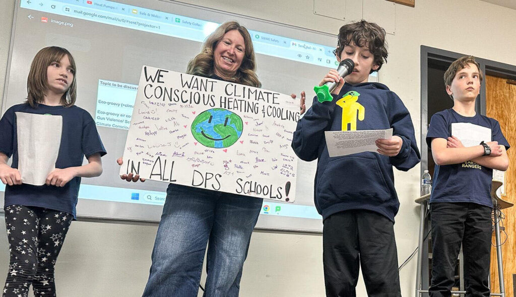 A young boy holds a microphone to his mouth as two other students and a teacher look on. The teacher holds a poster board with a drawing of Earth that says "We want climate conscious heating & cooling in all DPS schools."