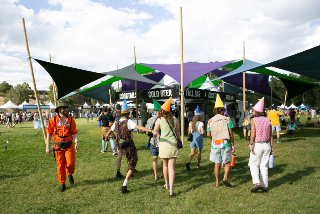A group of 7 people in pointy elfish hats frolic in a field, walking towards the "cold beer" and "cocktail" tent near the main stage.