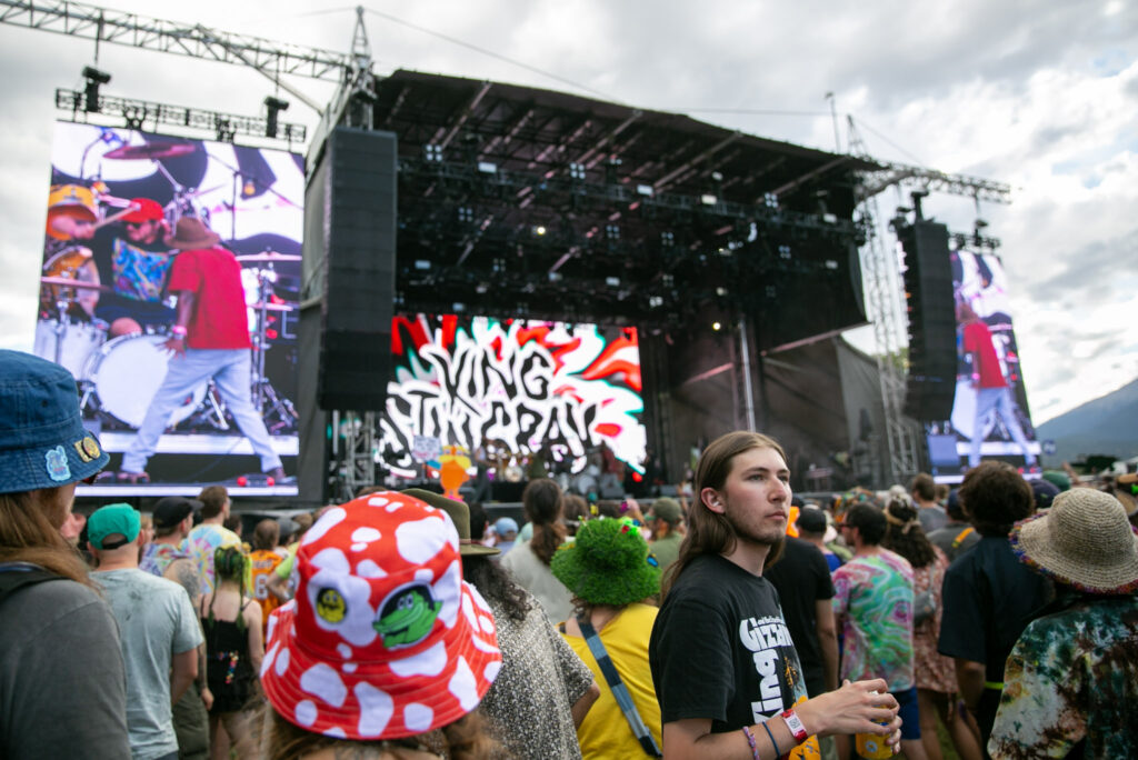 A stage with a big, "King Stingray" backdrop is surrounded by hundreds of fans. In the foreground, a man faces away from the stage, staring into the distance, as everyone around him looks towards the music.