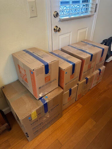 Image shows cardboard boxes stacked against a wall and door with blue tape sealing the tops shut.