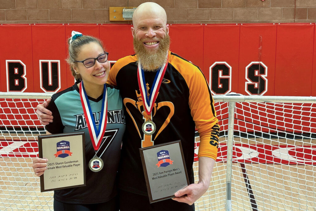 Two people stand with medals around their necks and holding plaques smiling in front of a goalball goal.