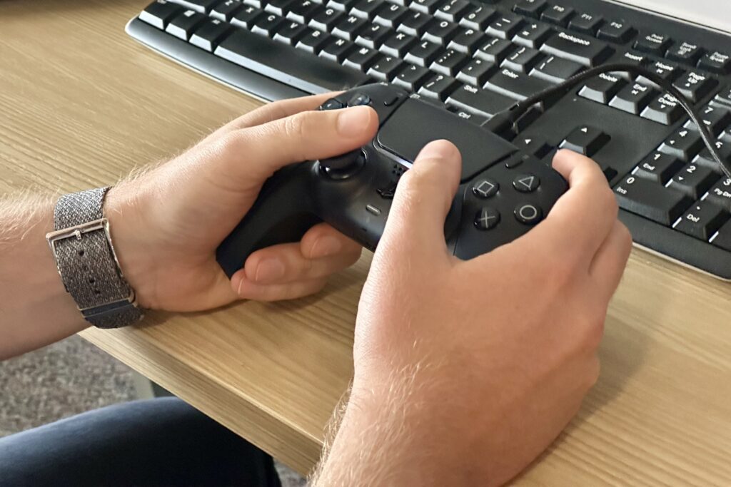 This a closeup of a person's hands holding a Playstation 5 controller in front of a keyhboard