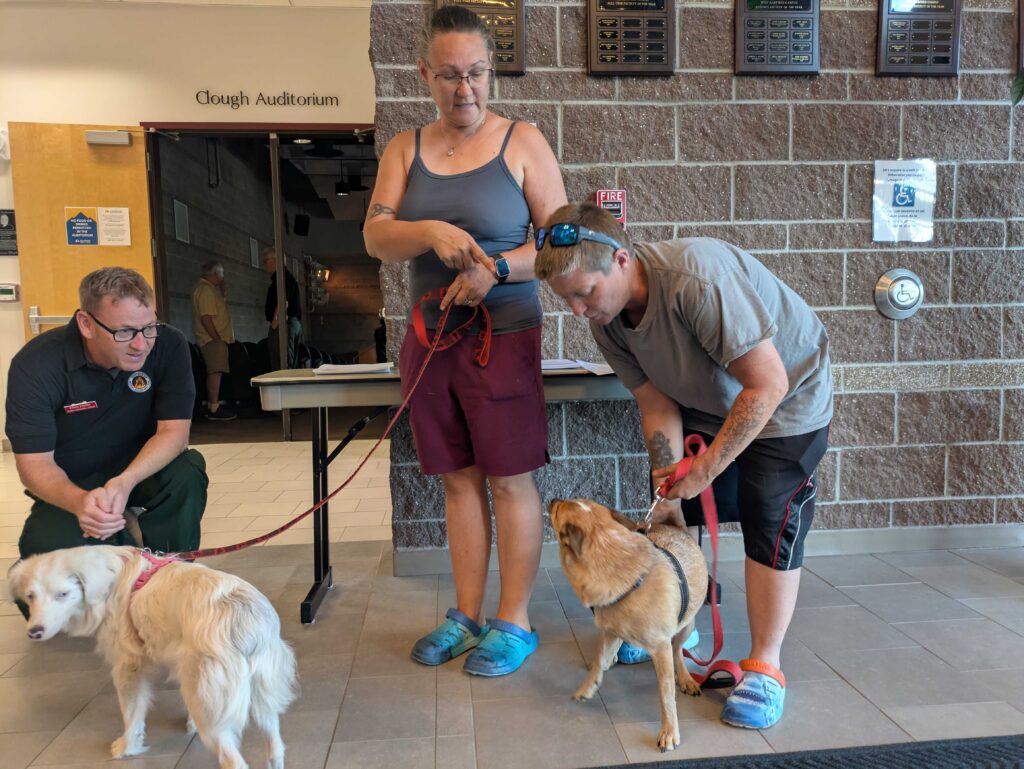 Two people stand with their dogs on leashes inside a building.