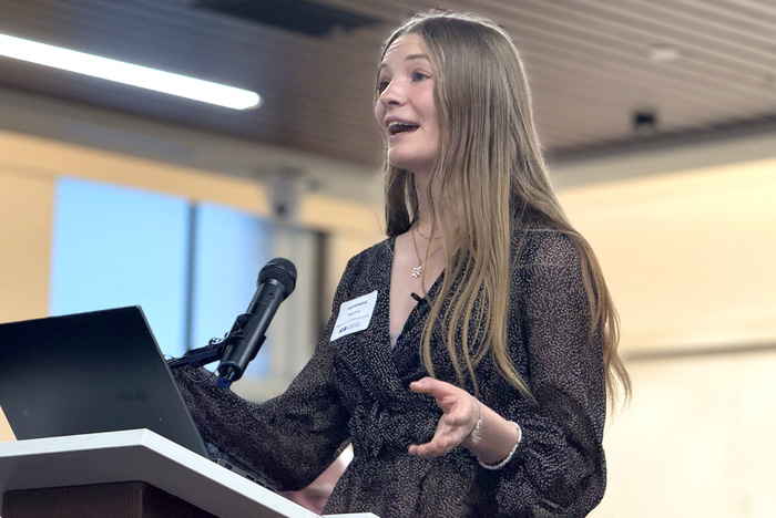 A young girl with long blonde hair speaks at a lecturn and makes a hand gesture while she speaks.