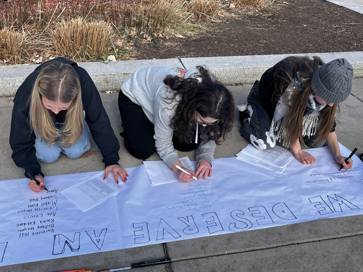 Three young women kneel on the sidewalk as they write messages on a banner.