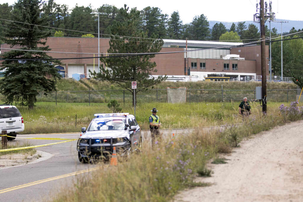 A police car with flashing lights, two officers, and yellow tape block a rural road near a brick building surrounded by trees and grassy areas.
