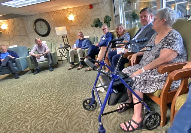 On the right of the frame, an older woman sits with a walker in front of her. She is speaking to a roomfull of people with six other people sitting in the round.