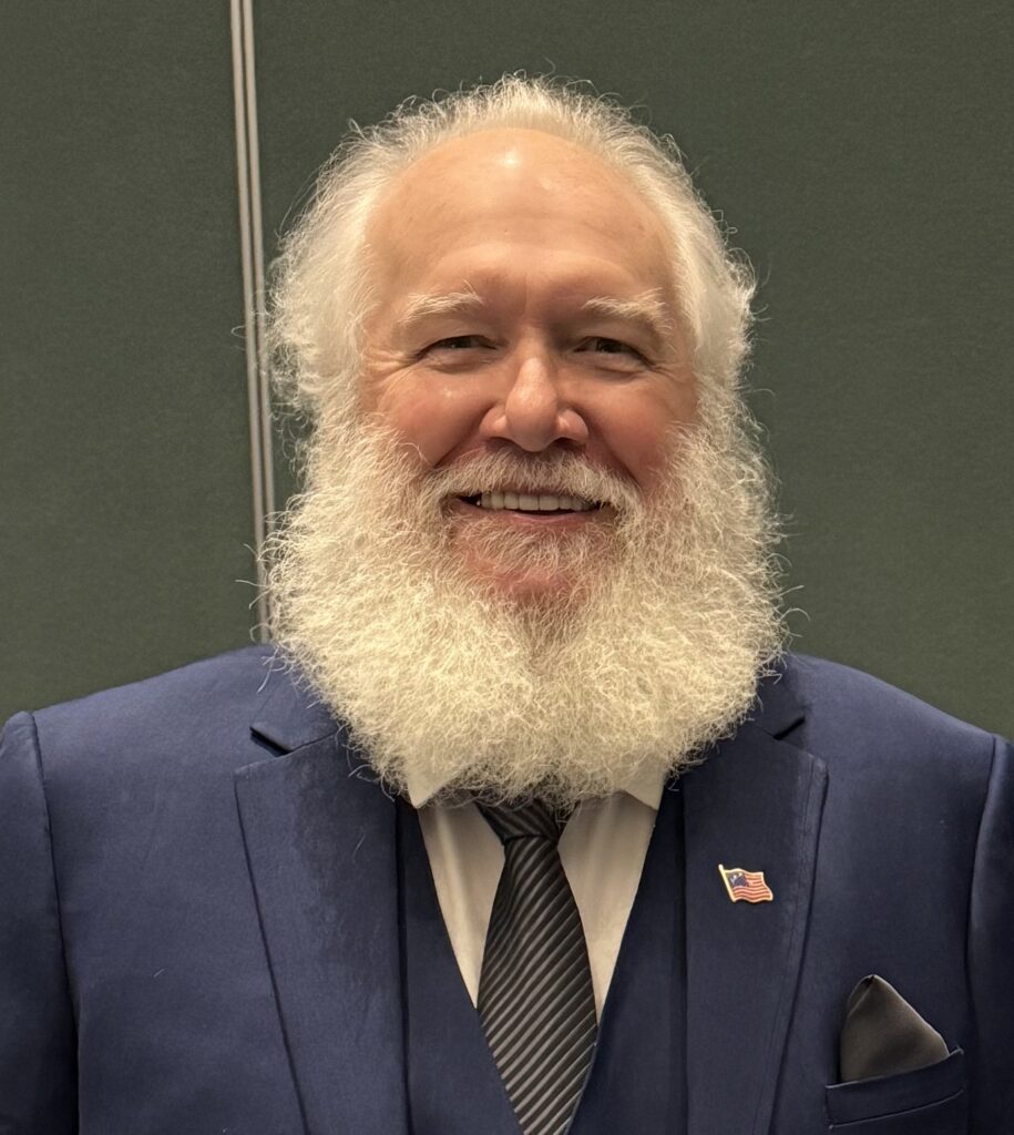 man wearing a blue suit and tie. He is smiling and has white hair and beard.