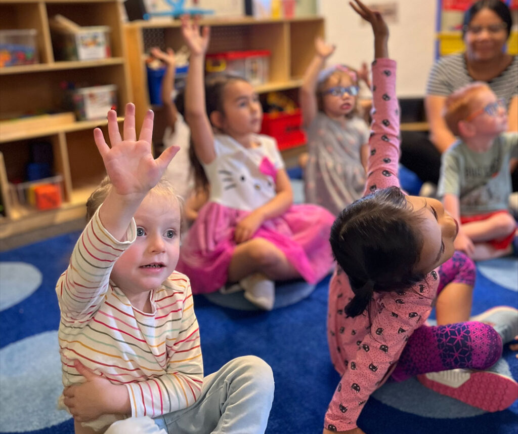A group of children sitting on the floor, raising their hands in the air. The children are of various ages and are spread out across the room, with some sitting closer to the front and others further back. The scene is focused on the children's interaction and participation in the activity.