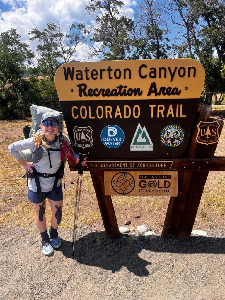 A woman in hiking gear stands in front of a sign that reads Waterton Canyon and Colorado Trail