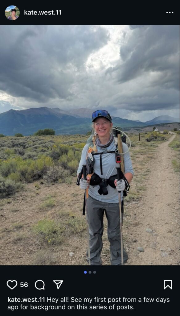 A woman in hiking gear stands on a trail with clouds and mountains in the background.