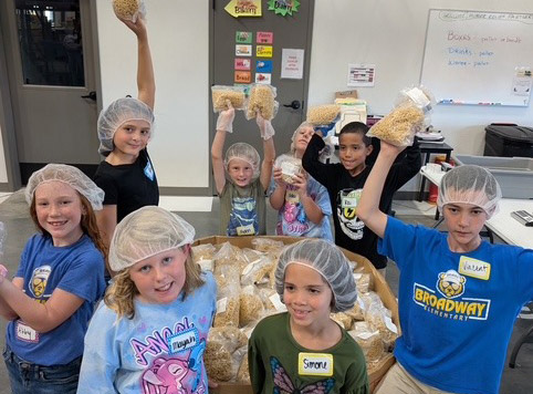 A group of children are standing in front of a box filled with food. They are posing for a picture, with some of them holding the box. The children are wearing hairnets.
