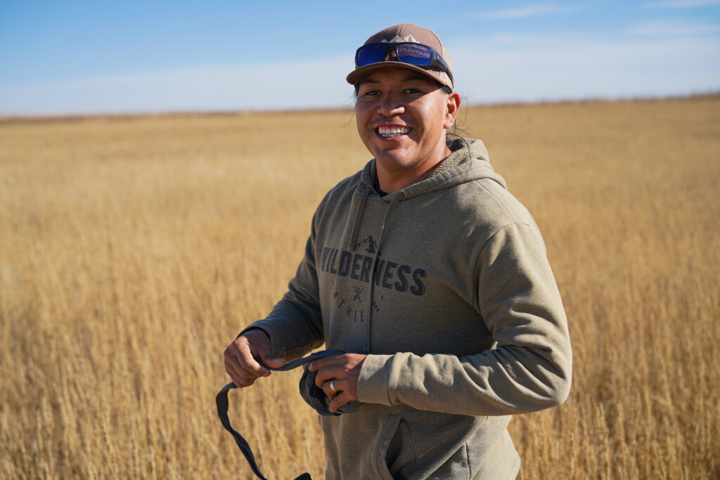 A man wearing a brown hat and a grey hoodie stands in a field covered with dry grass.. He is smiling and holding something in his hand.