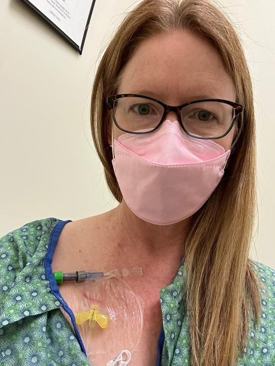 A woman in a pink mask and hospital gown with a tube sticking out of her looks into the camera.