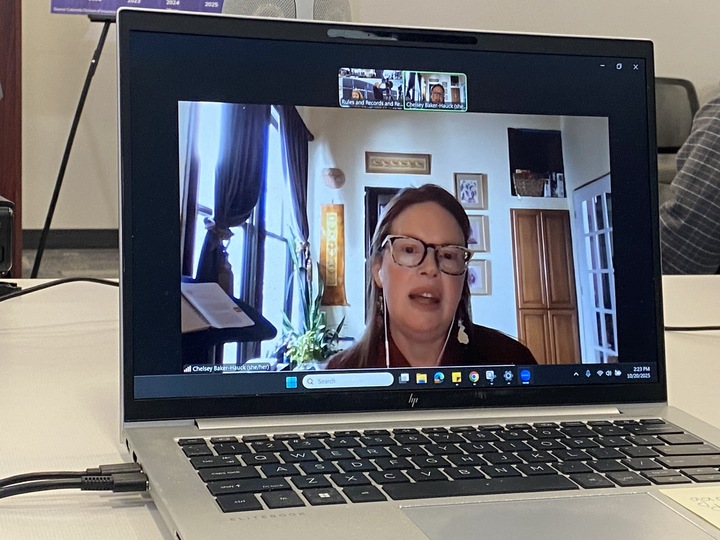 A laptop screen displays a woman talking on a video call. She is wearing glasses and has a headset on. The laptop is placed on a table.