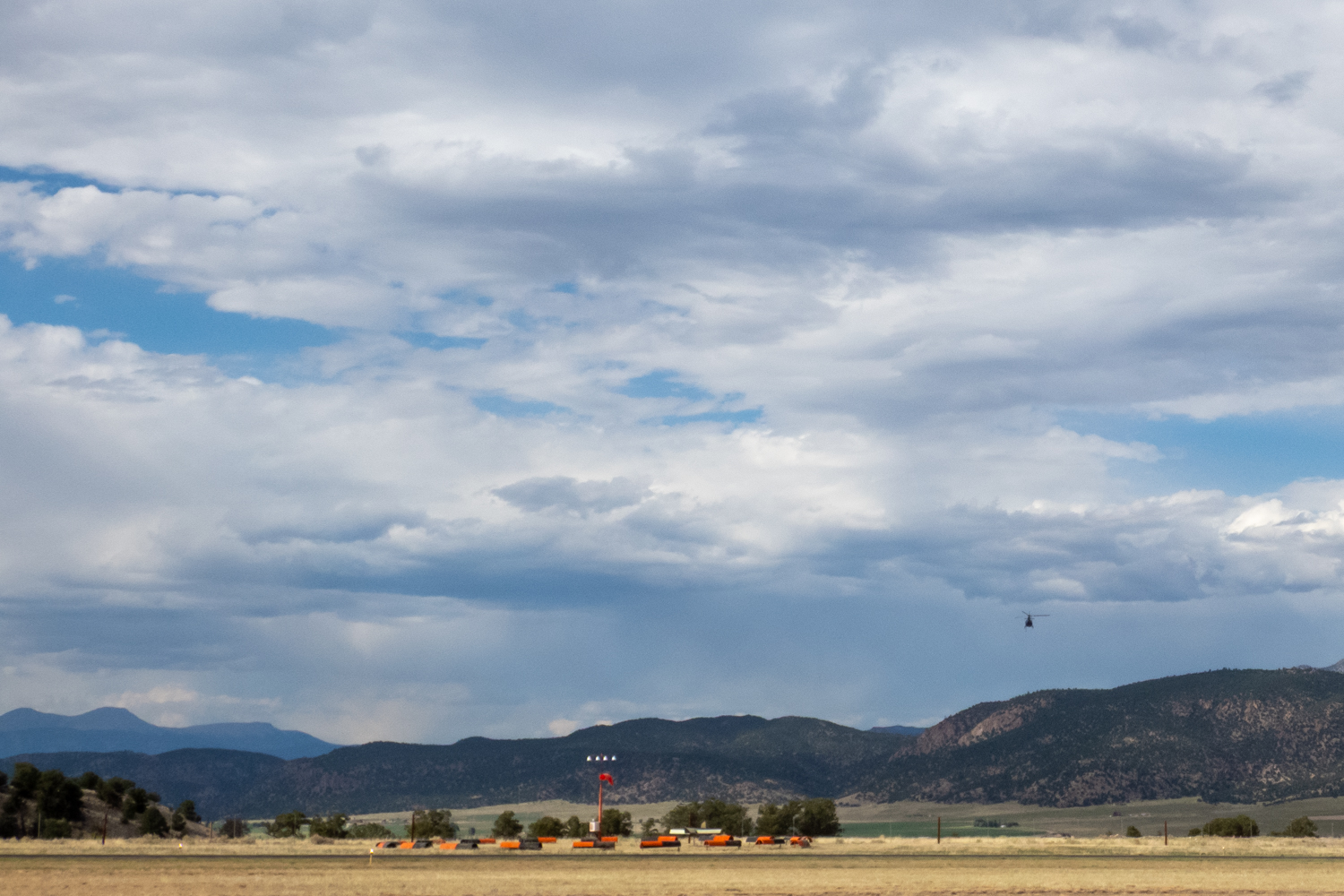A helicopter flies some distance away over a large valley with a group of orange airport equipment and lights. There are also a few trees in the background and mountains in the distance. The scene appears to be set in a rural or countryside location.