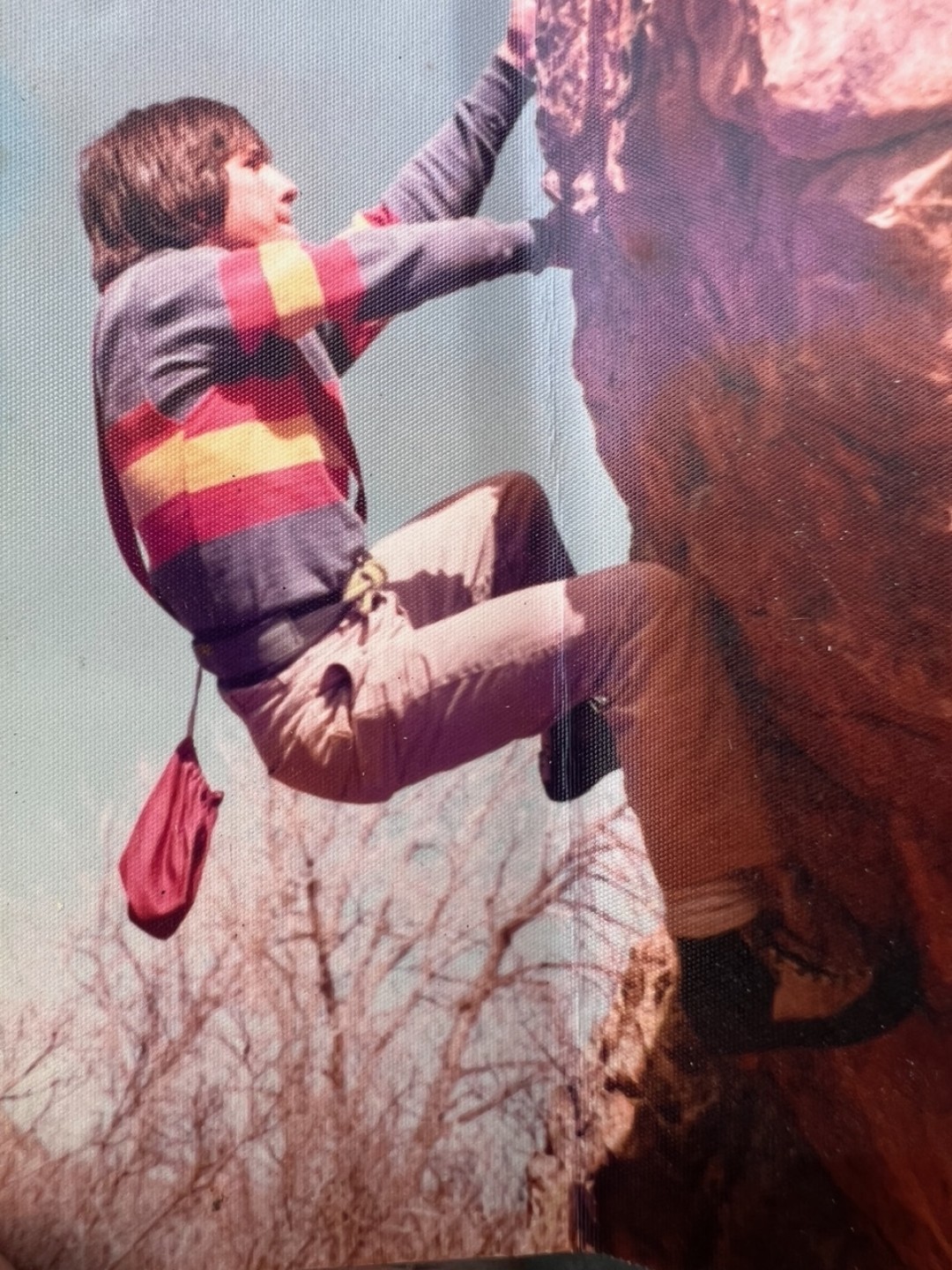 Image is a vintage photo circa 1978 showing a young man with a red, yellow, and blue striped shirt climbing a rockface in Garden of the Gods. His arms are outstretched and his legs are bent in a seated position.
