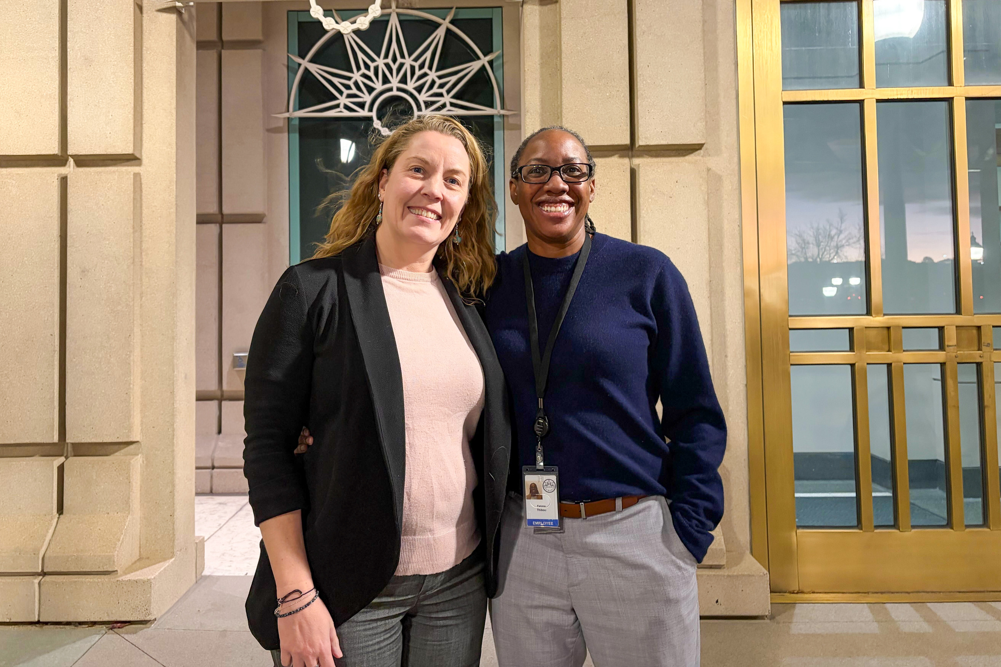 Two women are standing in front of a building, posing for a picture. One of them is wearing a black jacket and the other is wearing a blue sweater. They are both smiling.