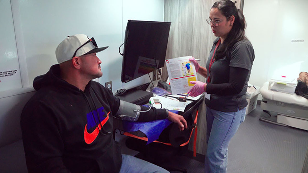 A man is sitting in a chair with a woman standing next to him. The woman is holding a piece of paper and pointing at it. The man is wearing a hat and has a blood pressure monitor on his arm.
